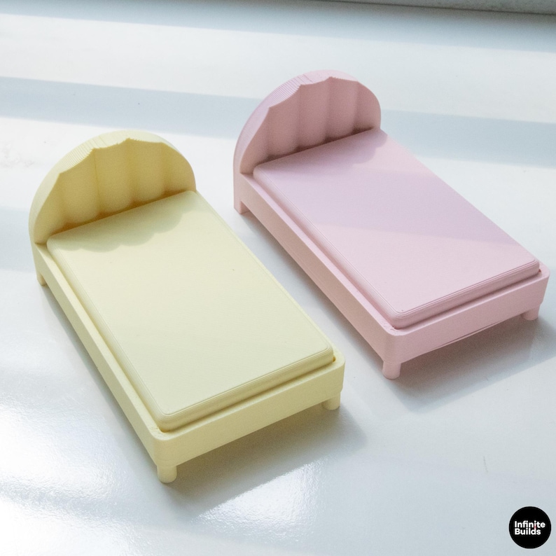 K&ouml;nnte beinhalten: Zwei Miniaturbetten, eines in blassem Gelb und eines in Rosa, werden gezeigt. Jedes Bett hat ein wellenf&ouml;rmiges Kopfteil, eine rechteckige Matratze und vier kleine Beine. Die Betten sind minimalistisch gestaltet.