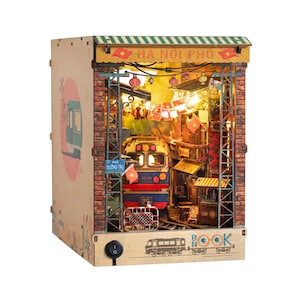 K&ouml;nnte beinhalten: Ein h&ouml;lzernes Buch-Eck mit einer Miniatur-Nachbildung einer vietnamesischen Stra&szlig;enszene mit einem Zug, Backsteinmauern und dem Text "HA NOI PHO". Die Szene wird von warmen Lichtern beleuchtet und enth&auml;lt Miniaturflaggen und Laternen.