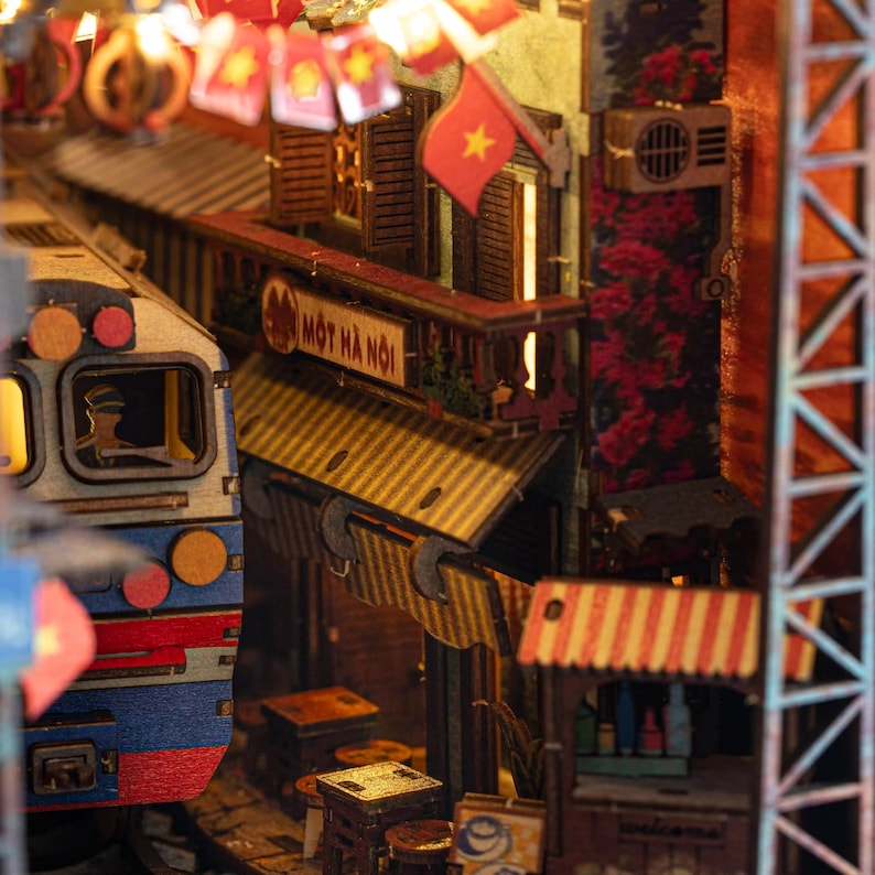 K&ouml;nnte beinhalten: Detailliertes Holzmodell einer vietnamesischen Stra&szlig;enszene mit einem Zug, Gesch&auml;ften und einer Flagge mit gelbem Stern. Das Modell enth&auml;lt Miniaturdetails wie Markisen, Schilder und florale Akzente.