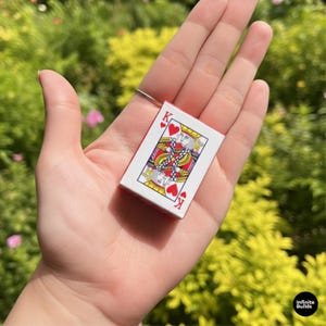 May include: A miniature playing card featuring the King of Hearts, held in a hand. The card has a red border and a detailed illustration of the king, with the suit symbols visible. The card is small, likely a miniature version.
