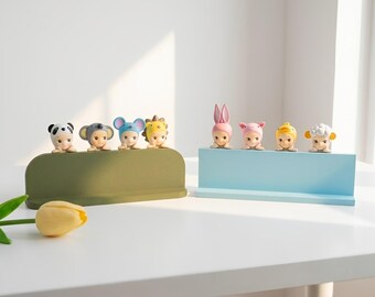 May include: Eight small figurines with animal-themed headgear arranged on two rectangular stands. The olive green stand holds panda, koala, mouse, and lion figures. The light blue stand holds rabbit, pig, chick, and sheep figures.