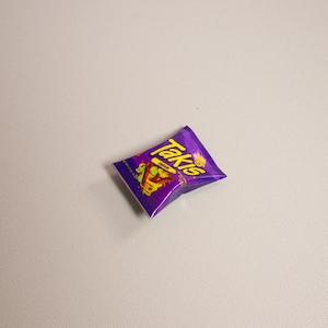 May include: A single, unopened bag of Takis Fuego tortilla chips. The bag is a vibrant purple with yellow and red text and graphics. The chips are a popular snack food.