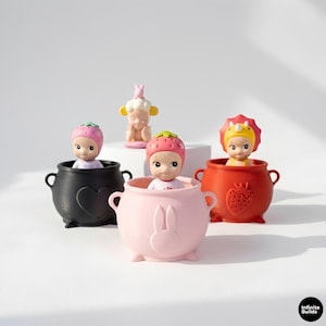 Puede incluir: Una colección de pequeñas y caprichosas figuras. Cada figura está dentro de un recipiente en forma de caldero. Los recipientes son negros, rosas y rojos, con elementos decorativos como un corazón, un conejo y una fresa. Las figuras tienen sombreros únicos.