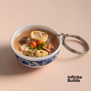 May include: A white ceramic bowl with a blue floral design filled with a miniature replica of a bowl of soup. The soup contains noodles, vegetables, and meat. The bowl is attached to a silver key chain.