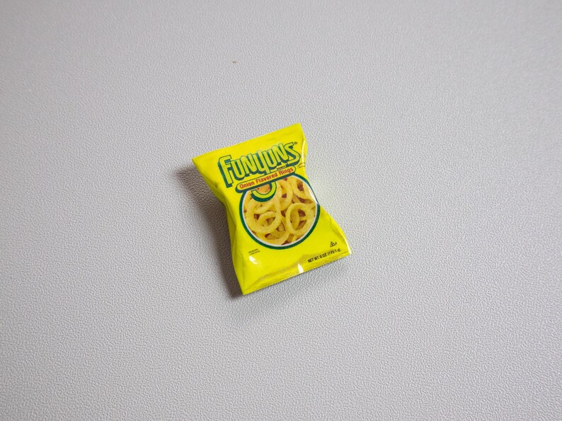 May include: A yellow bag of Funyuns Onion Flavored Rings snacks. The bag has the Funyuns logo in green and yellow, with an image of the onion rings. The bag is on a gray surface.