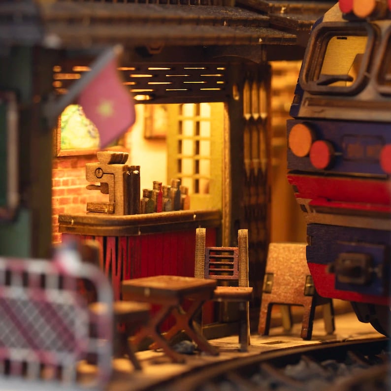 K&ouml;nnte beinhalten: Detailliertes Miniaturmodell einer Bahnhofsszene. Die Szene umfasst einen Zug, ein kleines Caf&eacute; mit Bar, Tischen und St&uuml;hlen. Eine Flagge ist sichtbar. Das Modell ist aus Holz gefertigt und hat eine warme, einladende Farbpalette.