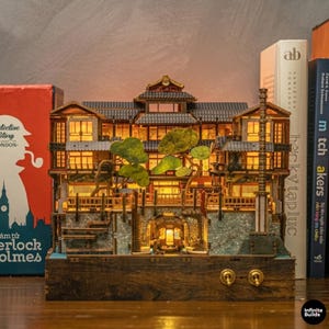 Könnte beinhalten: Detailliertes Holzmodell eines Gebäudes im japanischen Stil mit mehreren Ebenen, von innen beleuchtet. Das Modell weist komplizierte architektonische Details auf, darunter ein Dach im Pagodenstil, Balkone und Miniaturbäume. Die Basis besteht aus dunklem Holz.