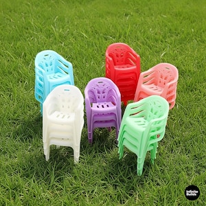 May include: A collection of colorful plastic armchairs in shades of red, pink, green, purple, white, and blue are stacked on a grassy surface. The chairs have a classic design with armrests and a slatted seat and back.