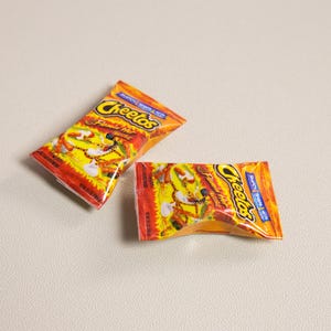 May include: Two small, orange and yellow Cheetos snack bags. The bags have the Cheetos logo and the words "Flamin' Hot" printed on them. The bags are on a light gray surface.