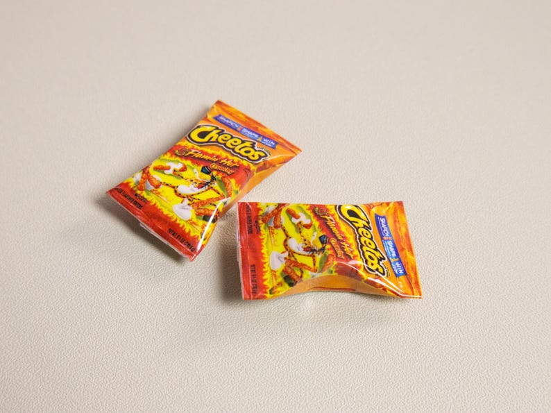 May include: Two small, orange and yellow Cheetos snack bags. The bags have the Cheetos logo and the words "Flamin' Hot" printed on them. The bags are on a light gray surface.