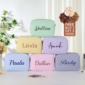 May include: Six striped fabric cosmetic bags with different names embroidered on them. The bags are in various colors, including light blue, yellow, pink, green, purple, and blue. The text "FREE BUY 1 GET 3" is visible in the top right corner of the image.