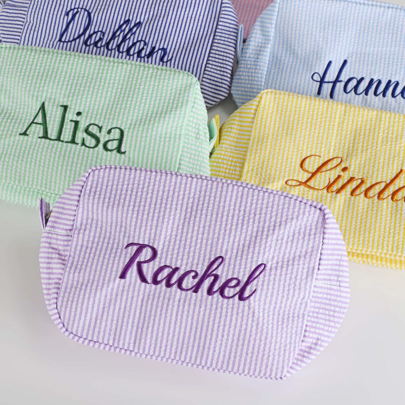 May include: Five personalized makeup bags in various colors, including purple, green, yellow, blue, and white. The bags are made of a striped fabric and have names embroidered on them, including "Alisa", "Dollar", "Hannah", "Linda", and "Rachel".