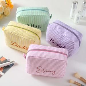 May include: Four personalized striped makeup bags in different colors. The bags are lined up in a row and have the names "Alisa", "Linda", "Rachel", and "Stacey" embroidered on them.