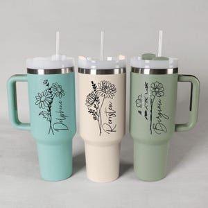 Könnte beinhalten: Drei isolierte Becher in Pastellfarben: Türkis, Creme und Salbeigrün. Jeder Becher hat ein Blumendesign und einen Namen in schwarzer Schrift: Delphine, Rensten und Virginia. Die Becher haben Griffe, Deckel und Strohhalme.