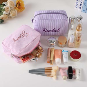 May include: Two pink and purple striped zippered makeup bags with the names "Alyssa" and "Rachel" embroidered on them. The bags are open and filled with various makeup products, including brushes, lipstick, foundation, and eyeshadow.