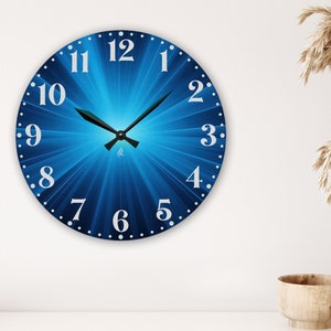 May include: Round wall clock with a blue radial design and white numerals. The clock hands are black. The clock is mounted on a white wall. A vase with dried plants and a wooden bowl are in the background.