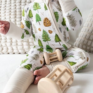 May include: A baby wearing a white and green long-sleeved onesie with a zip fastening and a forest print. The onesie has a white cuff on the sleeves and legs. The baby is sitting on a white bed with two wooden toys.