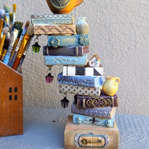 May include: A stack of decorative books with a succulent plant in a ceramic cup on top. The books are painted with various colors and patterns, including blue, purple, gold, and white. The books have decorative elements, such as a small house, a keyhole, and a lantern.