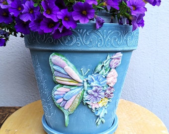 Puede incluir: Una maceta de cerámica azul con un diseño de mariposa y flores. La maceta está llena de petunias moradas.