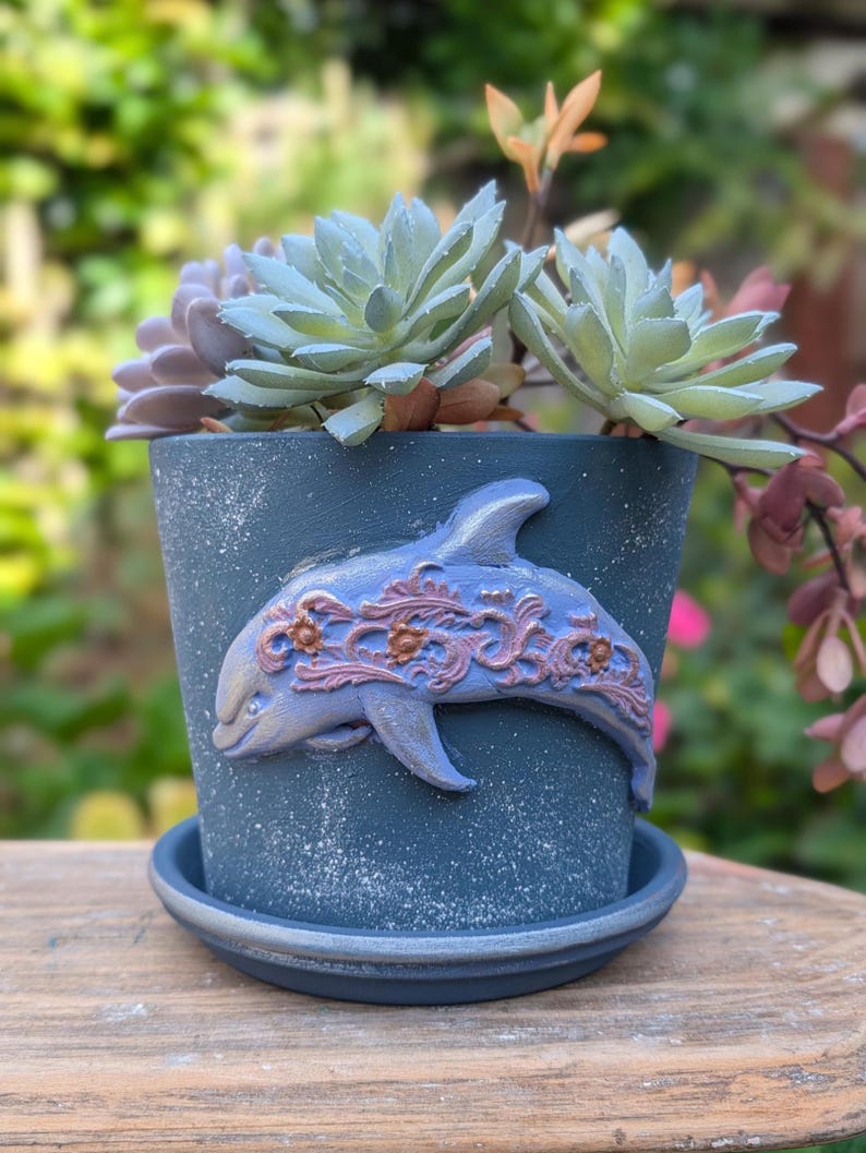 May include: A blue ceramic flower pot with a dolphin design and a matching saucer. The pot is filled with succulents in shades of green and purple. The dolphin is light blue with copper-colored accents. The pot sits on a wooden surface.