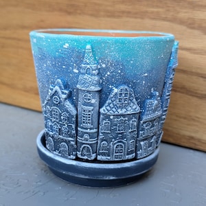 May include: A blue and white ceramic planter with a winter village scene. The planter has a snowy background and features a tall church building, houses, and a tower. The planter has a dark blue rim and a matching saucer.