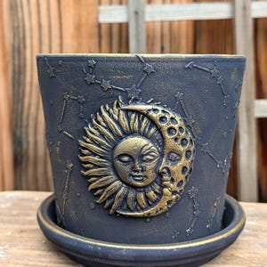 May include: A dark blue ceramic planter with a gold sun and moon design. The planter features constellations and a matching saucer. The sun and moon are embossed with detailed facial features. The planter is suitable for indoor or outdoor use.