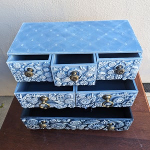 Puede incluir: Una c&oacute;moda de madera azul claro con cinco cajones. Cada caj&oacute;n tiene un dise&ntilde;o diferente con motivos florales y geom&eacute;tricos en relieve. El caj&oacute;n superior tiene un patr&oacute;n acolchado.