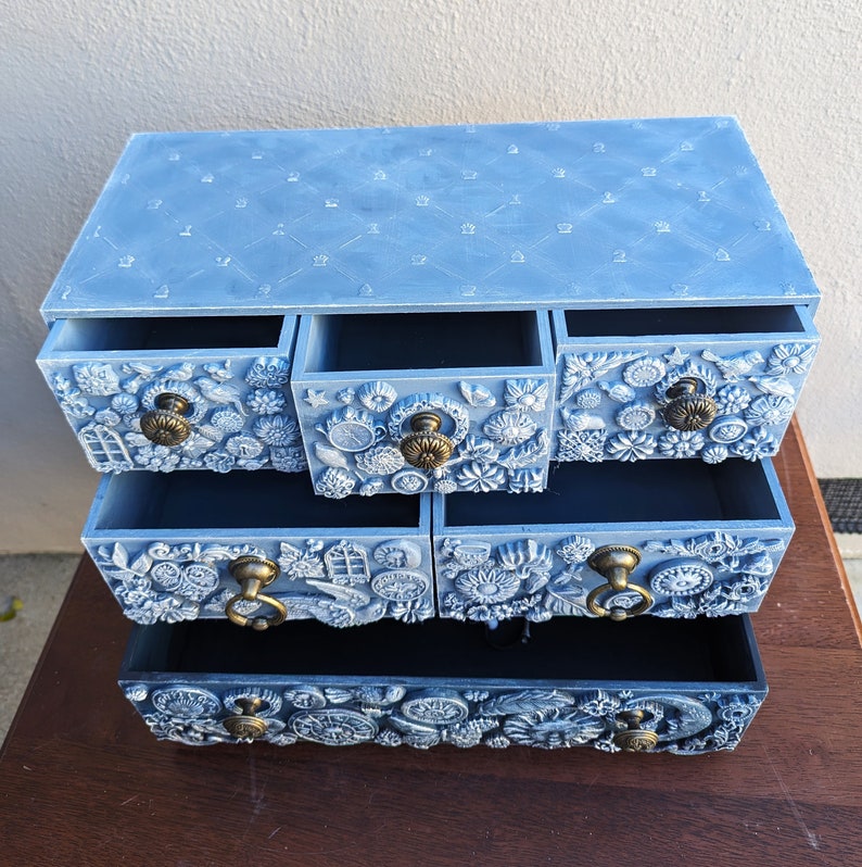 Puede incluir: Una c&oacute;moda de madera azul claro con cinco cajones. Cada caj&oacute;n tiene un dise&ntilde;o diferente con motivos florales y geom&eacute;tricos en relieve. El caj&oacute;n superior tiene un patr&oacute;n acolchado.