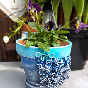 Pu&ograve; includere: Un vaso in ceramica blu e bianco con un design floreale decorativo. Il vaso &egrave; riempito con una piccola pianta con fiori viola.
