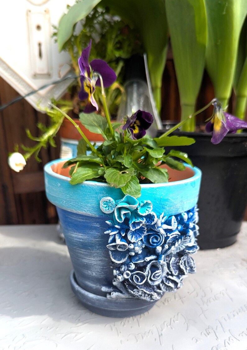 Pu&ograve; includere: Un vaso in ceramica blu e bianco con un design floreale decorativo. Il vaso &egrave; riempito con una piccola pianta con fiori viola.