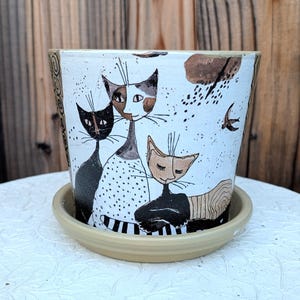 May include: A white ceramic plant pot with a matching saucer featuring a hand-painted illustration of three cats in black, white, and tan. The design includes abstract patterns, dots, and a small bird, with decorative spiral motifs on the side edges.