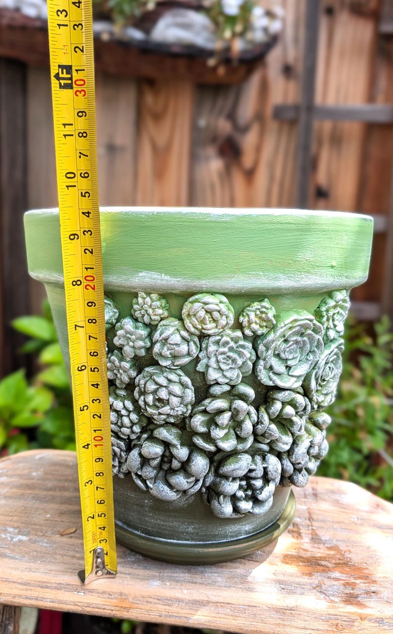 Op de afbeelding: Een groene keramische plantenpot, ongeveer 25 cm hoog, versierd met verhoogde succulentenontwerpen. De pot heeft een brede rand en een bijpassende schotel. De pot is geschilderd in een gedempte groene kleur.