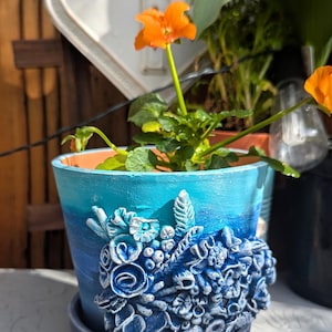 Pu&ograve; includere: Un vaso in ceramica blu e turchese con un design floreale 3D. Il vaso ha un design floreale blu e bianco con un bordo bianco. Il vaso &egrave; su una superficie bianca.