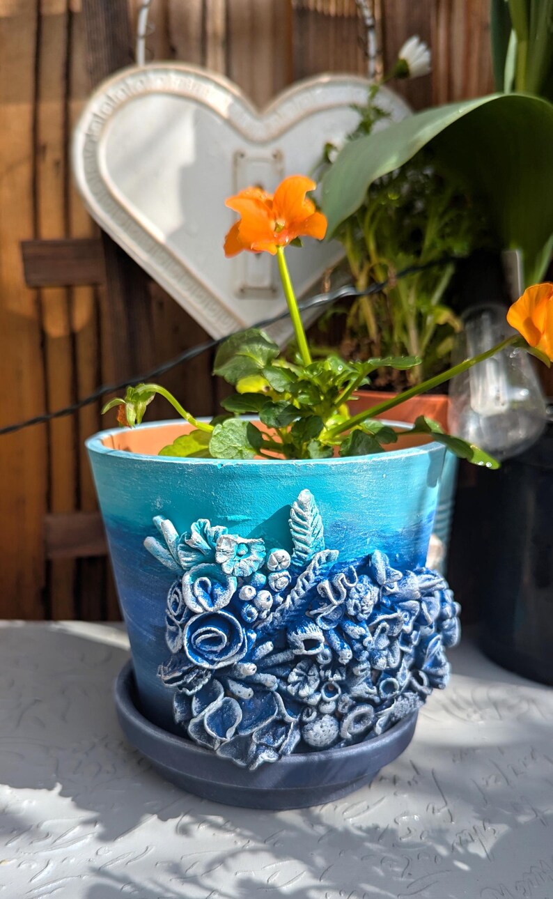Pu&ograve; includere: Un vaso in ceramica blu e turchese con un design floreale 3D. Il vaso ha un design floreale blu e bianco con un bordo bianco. Il vaso &egrave; su una superficie bianca.