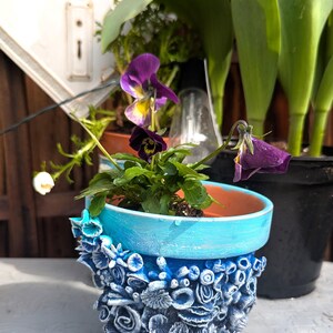 Pu&ograve; includere: Un vaso in ceramica blu e bianco con un design floreale testurizzato. Il vaso &egrave; riempito con una piccola pianta con fiori viola.