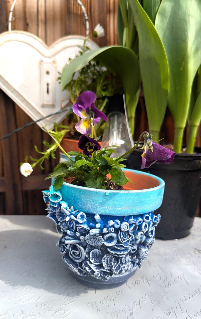 Pu&ograve; includere: Un vaso in ceramica blu e bianco con un design floreale testurizzato. Il vaso &egrave; riempito con una piccola pianta con fiori viola.