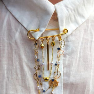 May include: A gold-toned safety pin brooch featuring multiple dangling chains adorned with small gold stars, crescent moons, white beads, and tiny blue square beads. A central vertical gold bar hangs among the delicate layered chains.