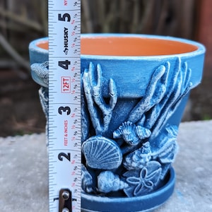 May include: A blue ceramic flower pot with a detailed underwater scene featuring seashells, coral, and fish. The pot is approximately 4 inches tall. The interior is orange, and the exterior is a textured blue.