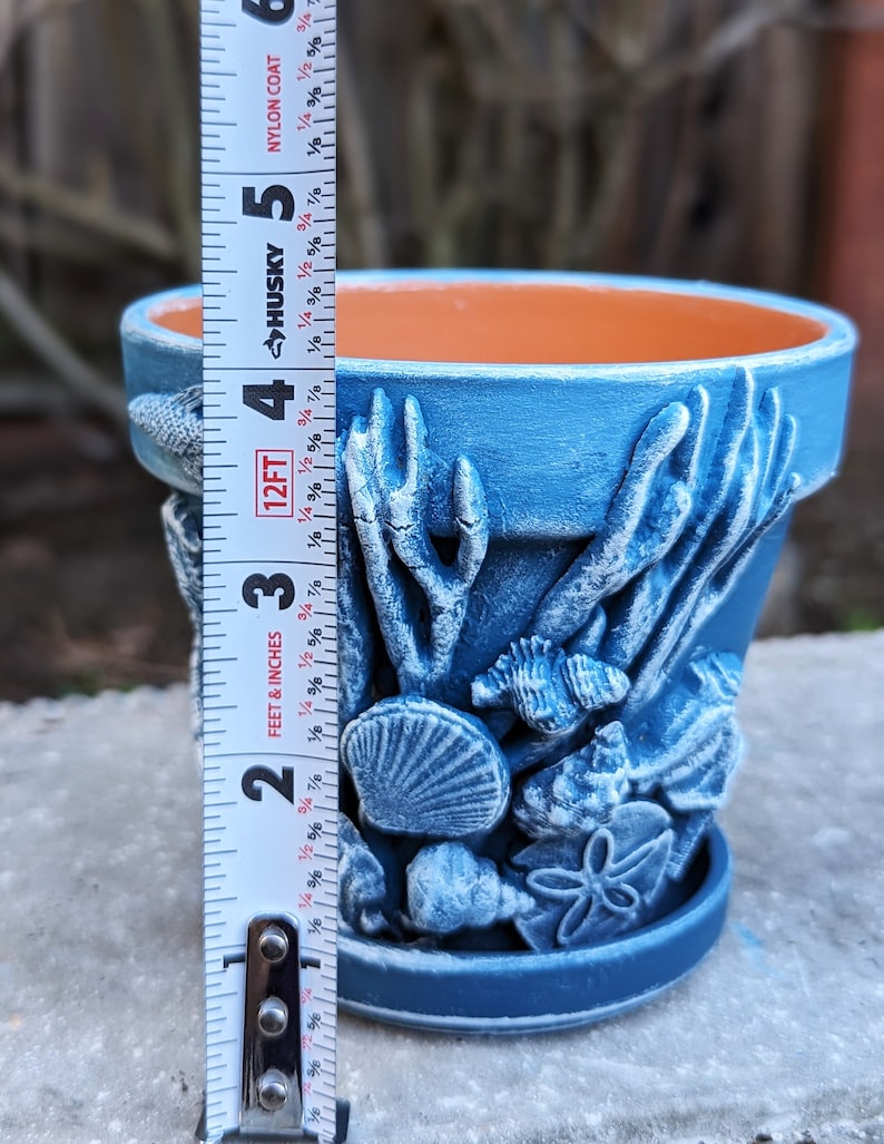 May include: A blue ceramic flower pot with a detailed underwater scene featuring seashells, coral, and fish. The pot is approximately 4 inches tall. The interior is orange, and the exterior is a textured blue.