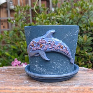 May include: A blue ceramic flower pot with a matching saucer. The pot features a raised dolphin design with ornate detailing in shades of lavender and bronze. The pot has a speckled finish and is suitable for indoor or outdoor use.