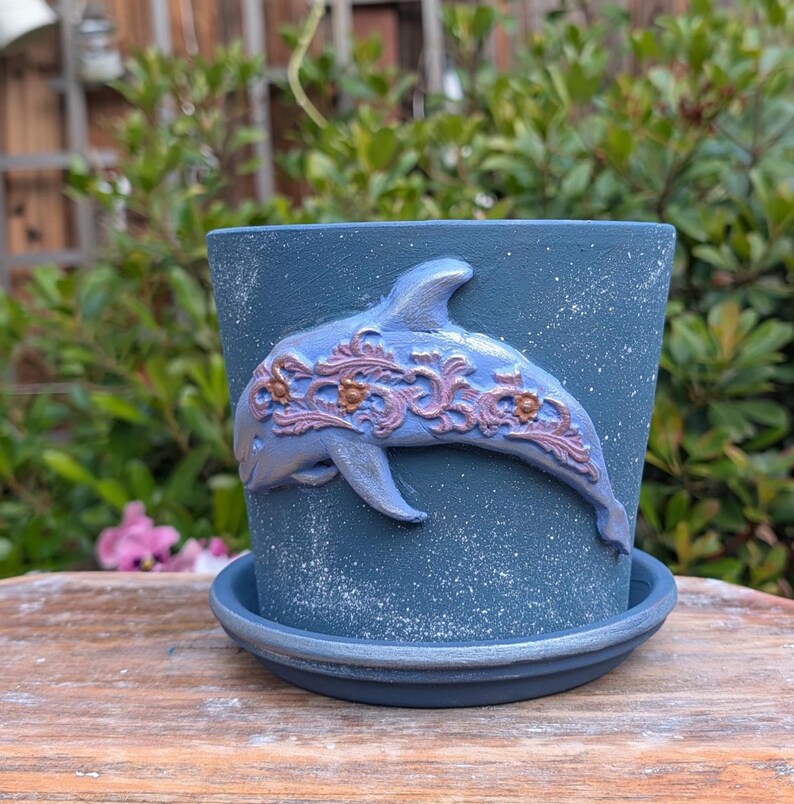 May include: A blue ceramic flower pot with a matching saucer. The pot features a raised dolphin design with ornate detailing in shades of lavender and bronze. The pot has a speckled finish and is suitable for indoor or outdoor use.
