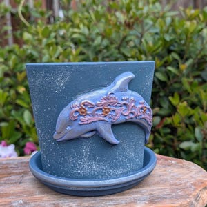 May include: A blue ceramic flower pot with a matching saucer. The pot features a raised dolphin design with pink and gold accents. The pot has a textured, speckled finish and is suitable for indoor or outdoor use.