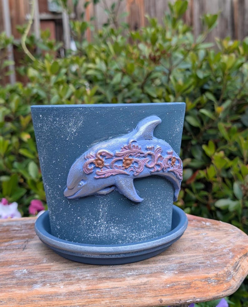 May include: A blue ceramic flower pot with a matching saucer. The pot features a raised dolphin design with pink and gold accents. The pot has a textured, speckled finish and is suitable for indoor or outdoor use.