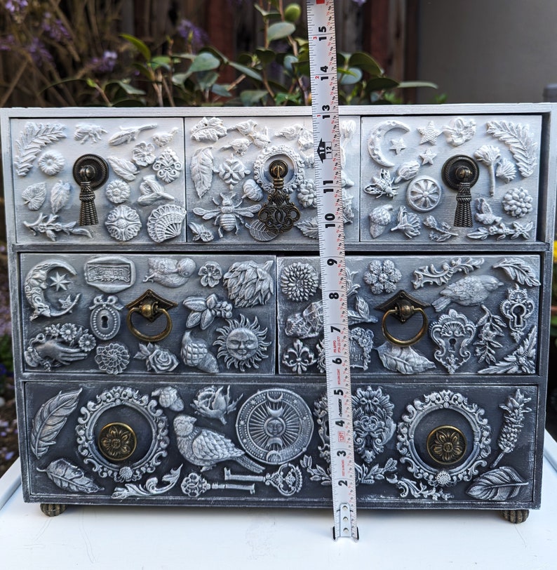 Puede incluir: Una c&oacute;moda de madera blanca con tres cajones. Cada caj&oacute;n est&aacute; decorado con intrincados tallados de flores, hojas y otros dise&ntilde;os. La c&oacute;moda est&aacute; pintada en un acabado blanco desgastado, lo que le da un aspecto vintage.