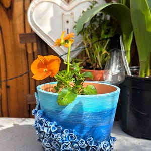 Pu&ograve; includere: Un vaso in ceramica blu e bianco con un design testurizzato a forma di conchiglia. Il vaso &egrave; riempito con una piccola pianta di fiori arancioni.