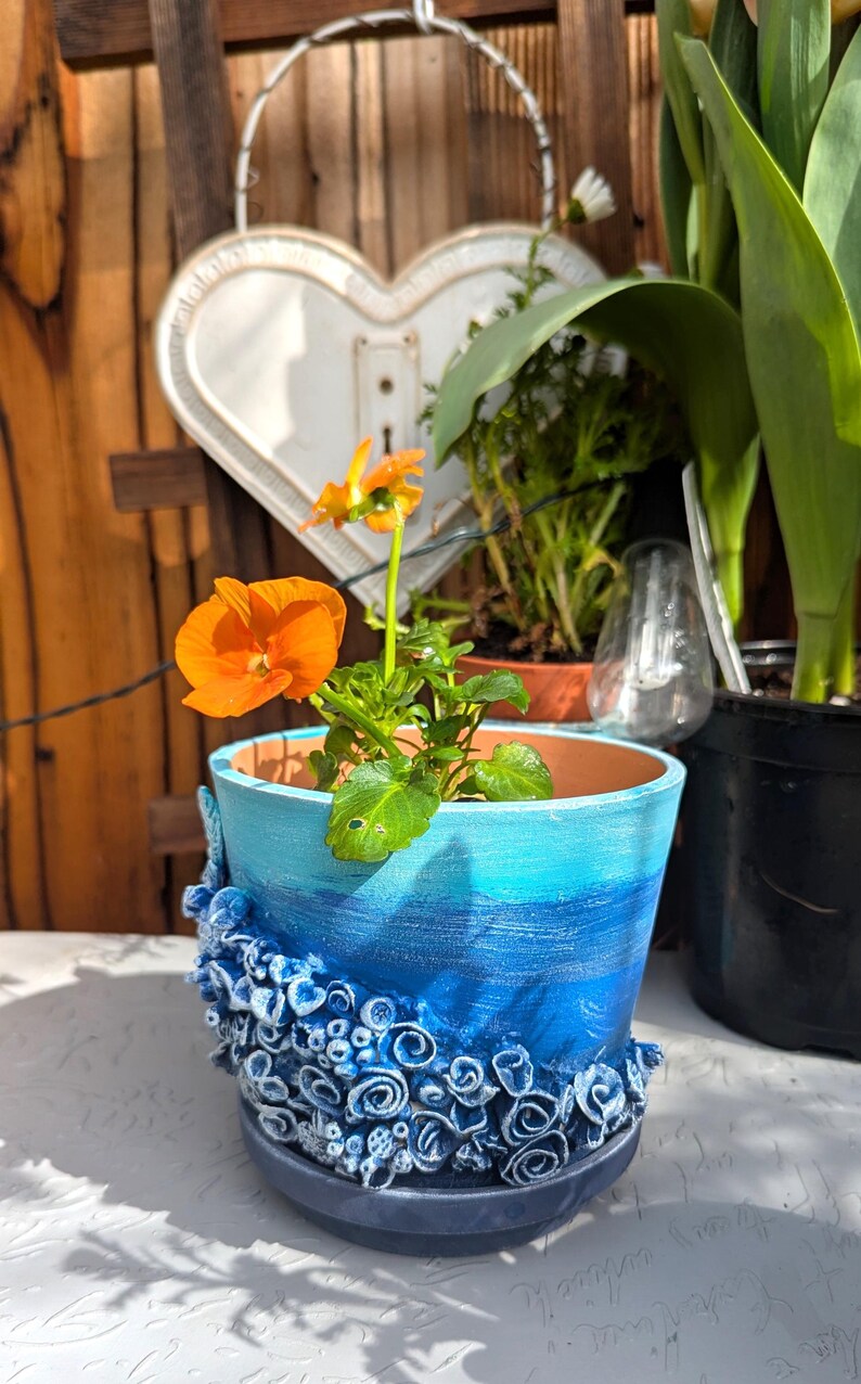 Pu&ograve; includere: Un vaso in ceramica blu e bianco con un design testurizzato a forma di conchiglia. Il vaso &egrave; riempito con una piccola pianta di fiori arancioni.
