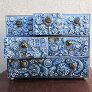 Puede incluir: Una caja de madera pintada de azul con cuatro cajones. Cada caj&oacute;n est&aacute; decorado con un dise&ntilde;o diferente de flores, hojas y otros elementos decorativos en relieve de metal. La caja tiene un acabado desgastado y est&aacute; adornada con pomos de metal dorados.