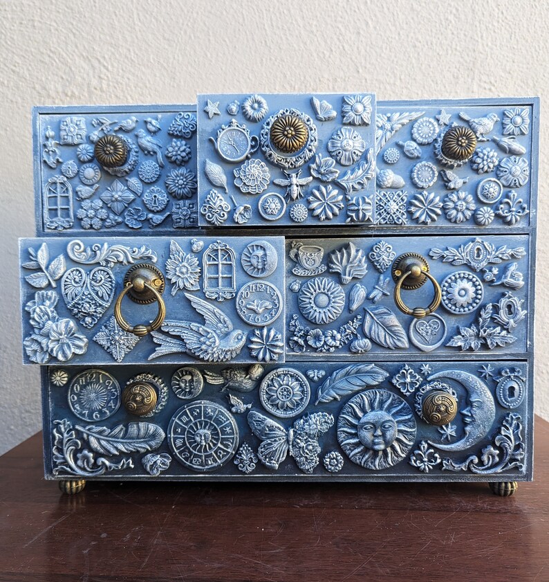 Puede incluir: Una caja de madera pintada de azul con cuatro cajones. Cada caj&oacute;n est&aacute; decorado con un dise&ntilde;o diferente de flores, hojas y otros elementos decorativos en relieve de metal. La caja tiene un acabado desgastado y est&aacute; adornada con pomos de metal dorados.