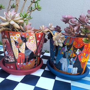 May include: Two decorative ceramic flower pots with cat illustrations. One pot is red with a floral and cat design, the other is blue with a cat design. Each pot holds a succulent plant and sits on a matching saucer.