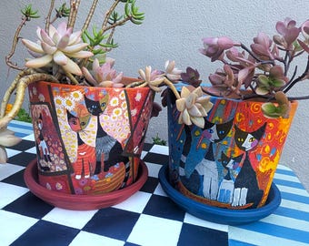 May include: Two ceramic plant pots with saucers, each featuring colourful painted cat illustrations. The left pot is orange with floral patterns and cats, while the right pot is blue and orange with a group of cats. Both contain live succulent plants.